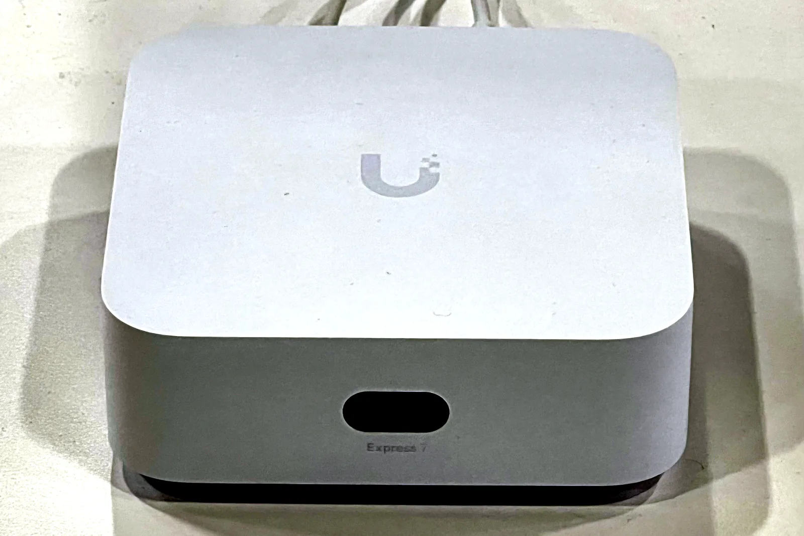 A white Ubiquiti UniFi Express 7 gateway seen from above and the front on a white surface, the Ubiquiti mark on its lid, its small status screen and the Express 7 name on the front edge