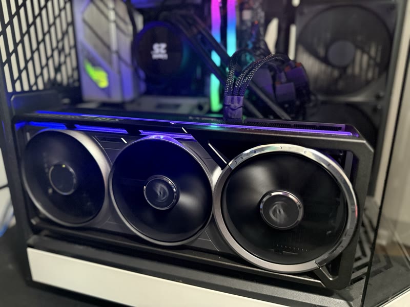 Close-up of the ASUS ROG Astral RTX 5080 OC quad-fan shroud: three exposed fans with center and offset ROG hubs, dial markings visible on the right-most fan, ARGB top bar lit purple, NZXT Kraken AIO display showing in background