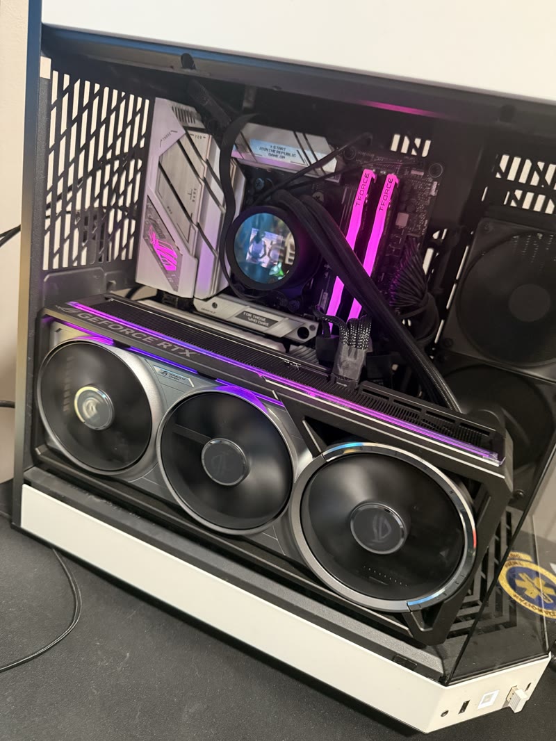 ASUS ROG Astral RTX 5080 OC installed in TechFuelHQ test bench: quad-fan cooler visible, ROG ARGB strip lit pink, T-Force RGB DDR5 modules behind, AIO pump cap visible above