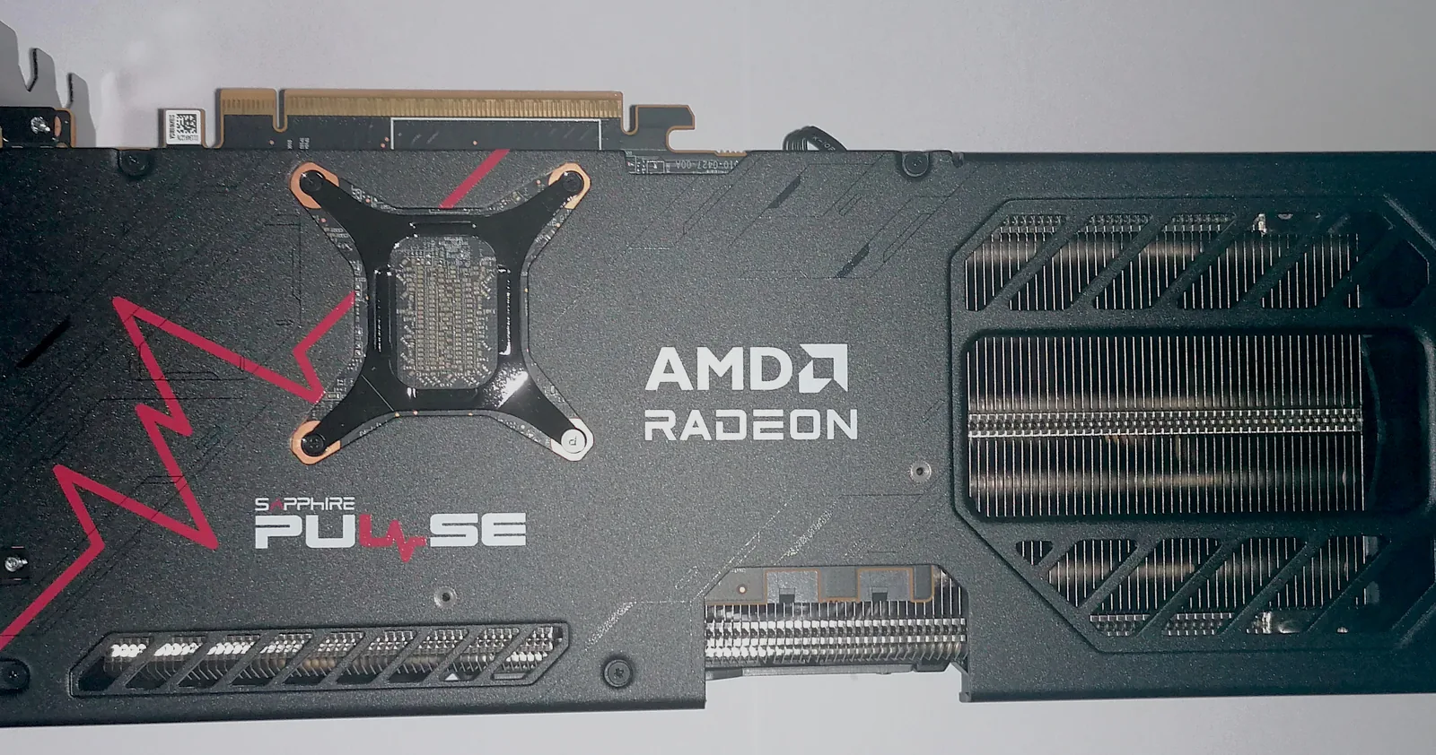 The backplate of a Sapphire Pulse Radeon RX 9070 XT graphics card on a white surface, AMD Radeon and Sapphire Pulse lettering on the dark plate, heatsink fins through the vent