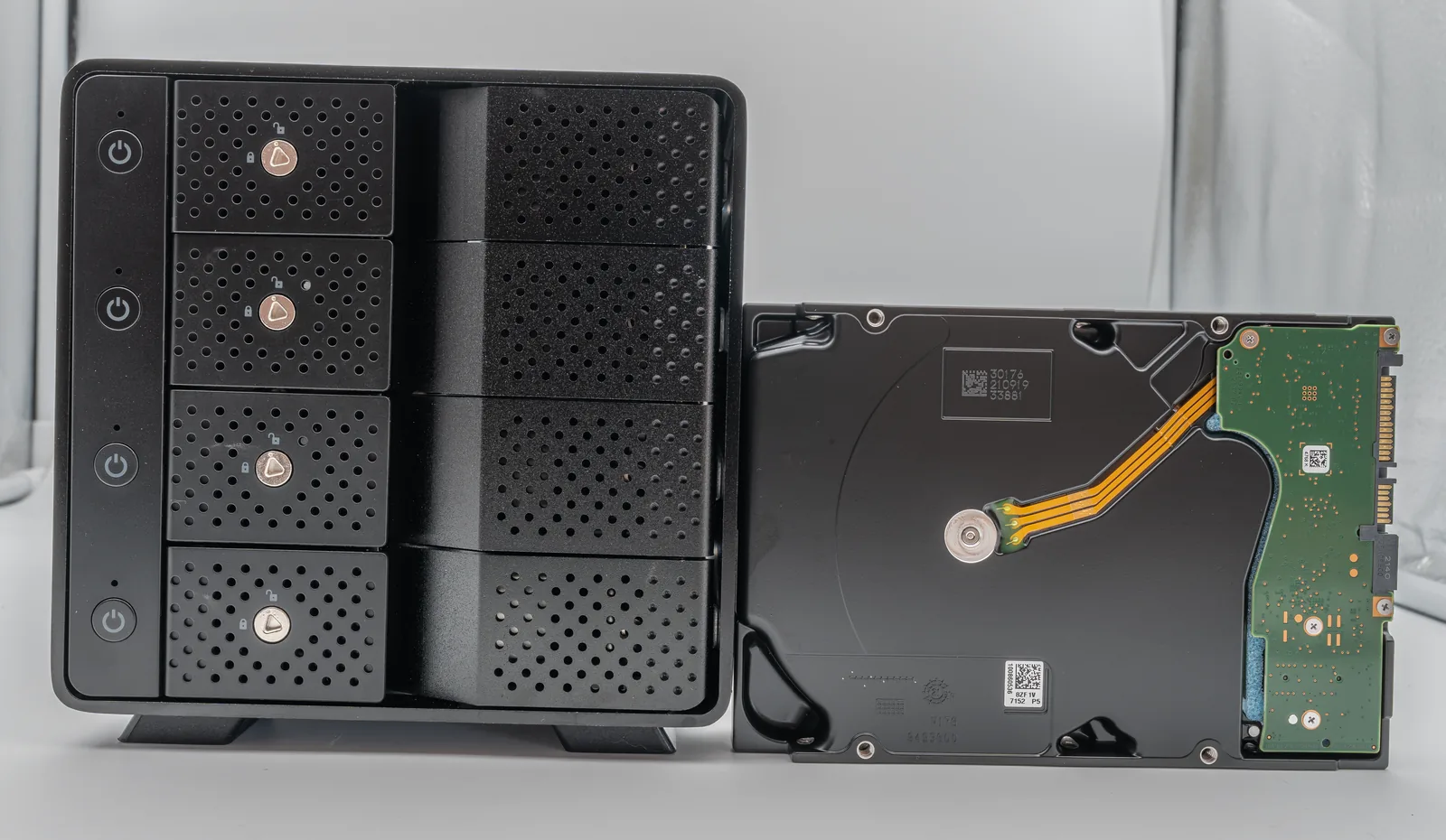 A black four-bay Sabrent direct-attached storage enclosure standing beside a bare 3.5-inch Seagate Exos hard drive on a white surface