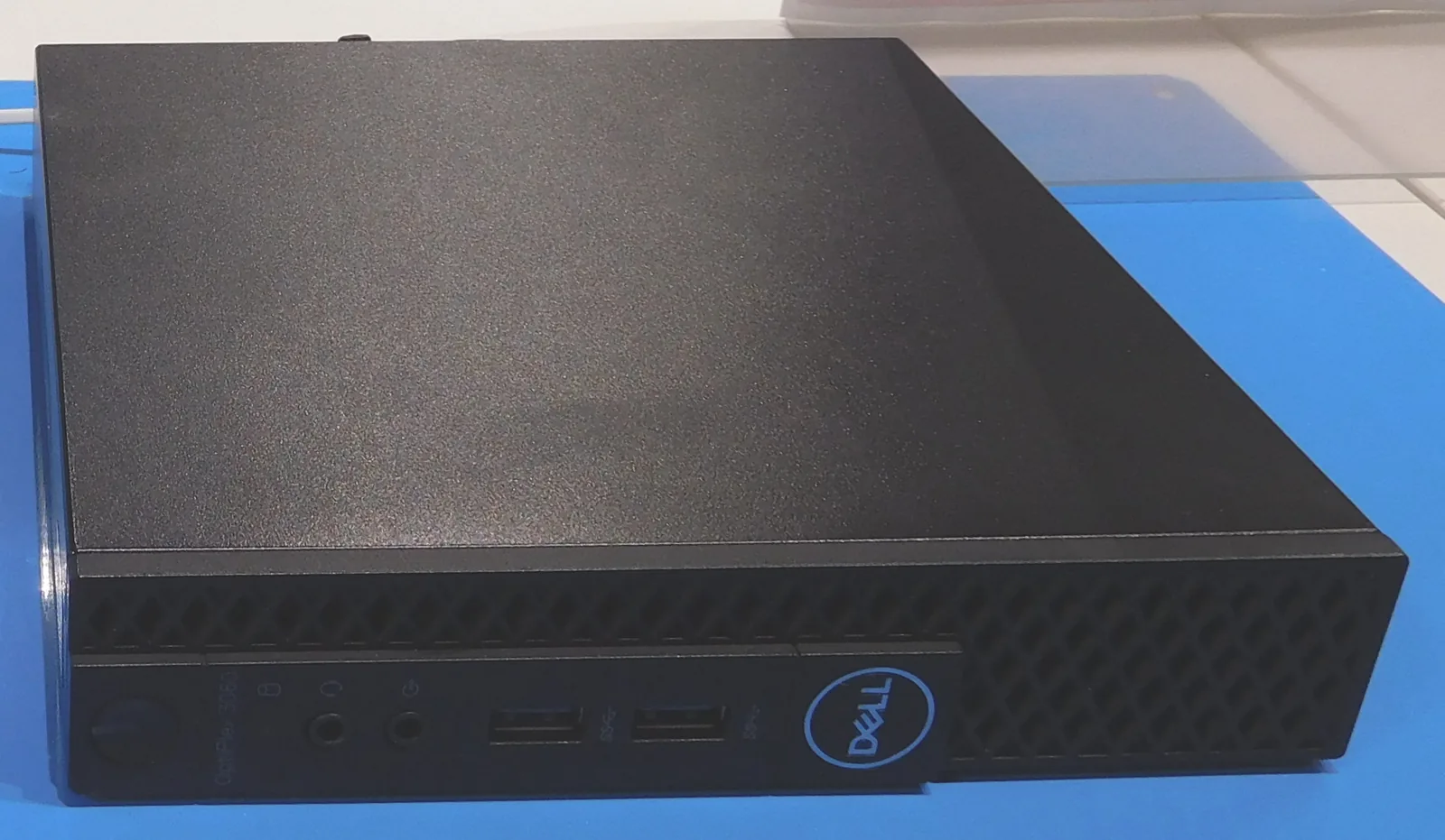 A black Dell OptiPlex 3060 Micro desktop lying flat on a blue cutting mat, its front USB ports and Dell badge facing the camera