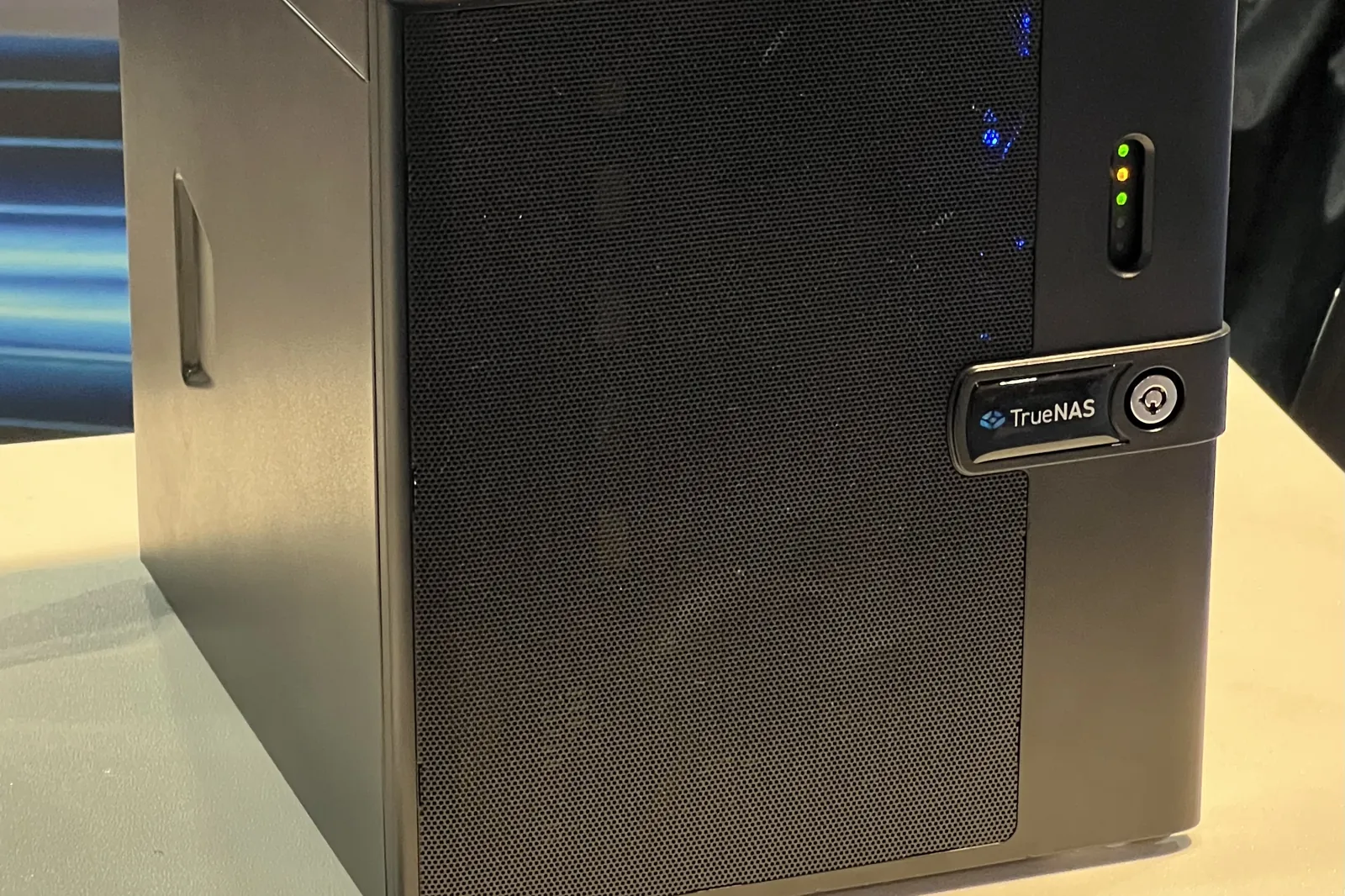 The front of a TrueNAS Mini X+ storage server, a black tower with a perforated drive door, status LEDs and the TrueNAS badge, on a show-floor table