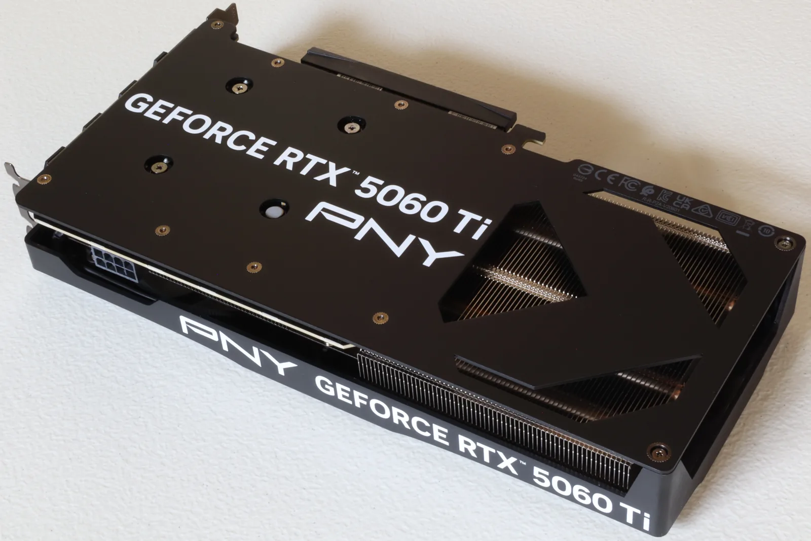 The backplate of a PNY GeForce RTX 5060 Ti 16GB graphics card on a white surface, GeForce RTX 5060 Ti and PNY lettering across the black plate, heatsink fins showing through the cut-out