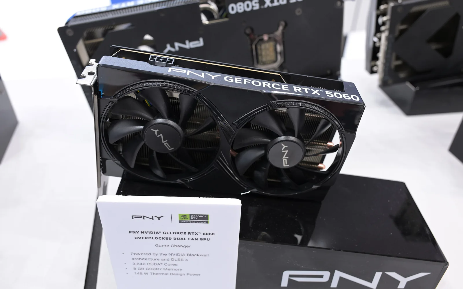 A PNY GeForce RTX 5060 dual-fan graphics card on a stand beside its retail box at a trade show display