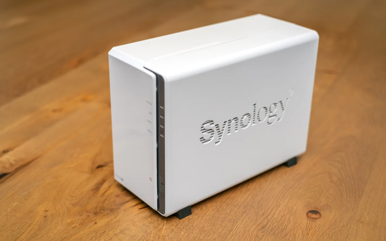 A white two-bay Synology DiskStation DS223J network-attached storage unit on a wooden desk, its drive-status lights along the front edge
